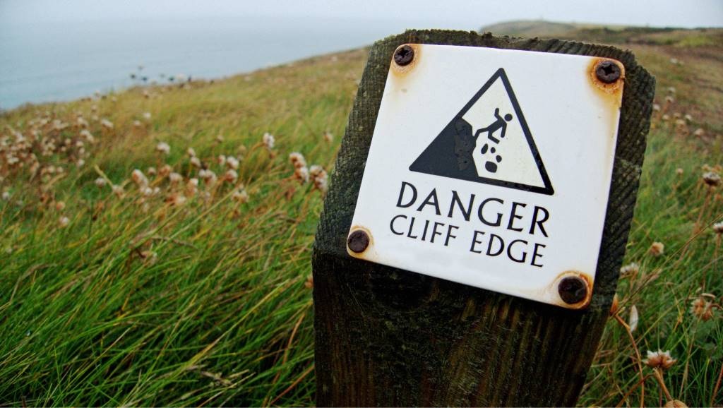 "Danger cliff edge" sign on a windswept grassy landscape. How does tourism avert disaster?