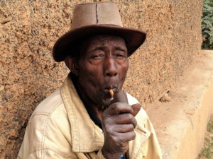 Rwandan man sits against a mud or clay wall smoking a pipe, looking at the camera. He's earing a wide=brimmed hat and a tan jacket.
